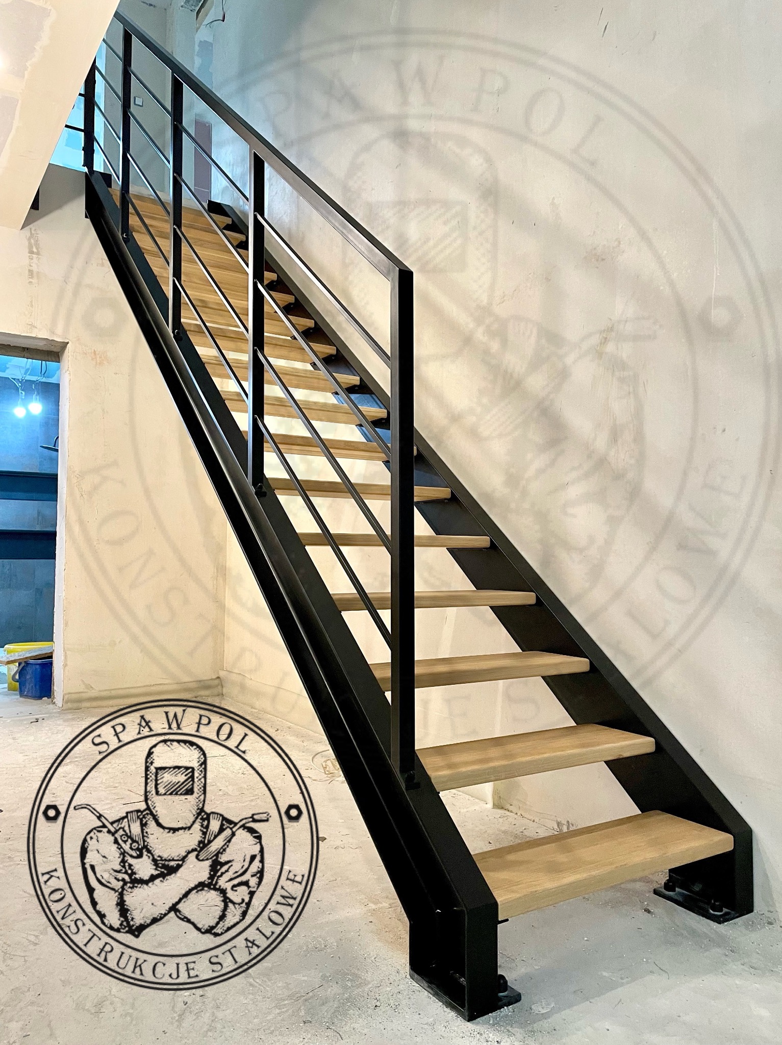 Nowoczesne schody metalowe z drewnianymi stopniami, czarna balustrada, widoczne logo firmy na ścianie i podłodze, surowy stan wykończenia wnętrza.