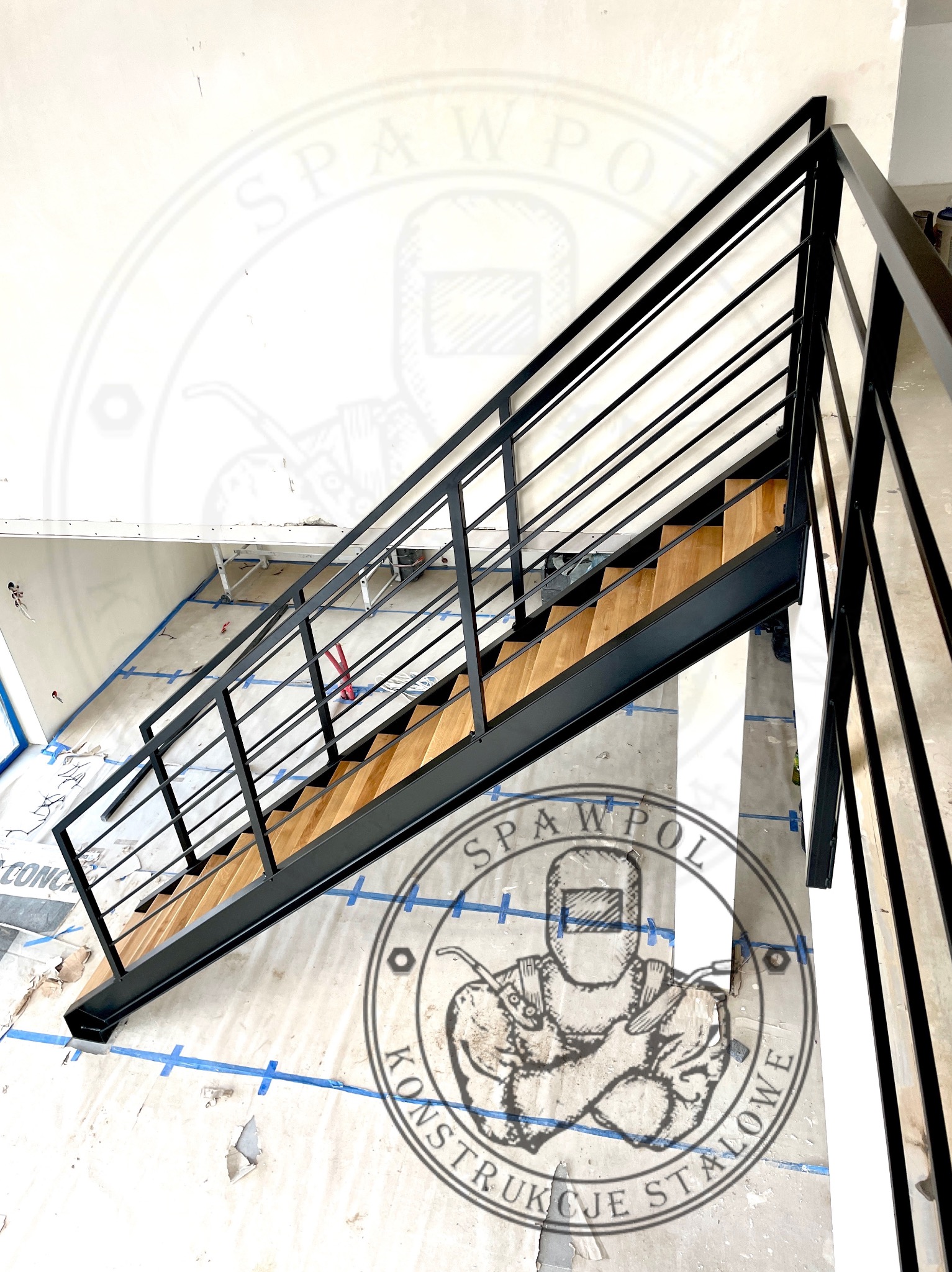 Nowoczesne schody metalowe z drewnianymi stopniami i czarną balustradą, widok z dołu, na podłodze zabezpieczenia budowlane, w tle logo firmy.