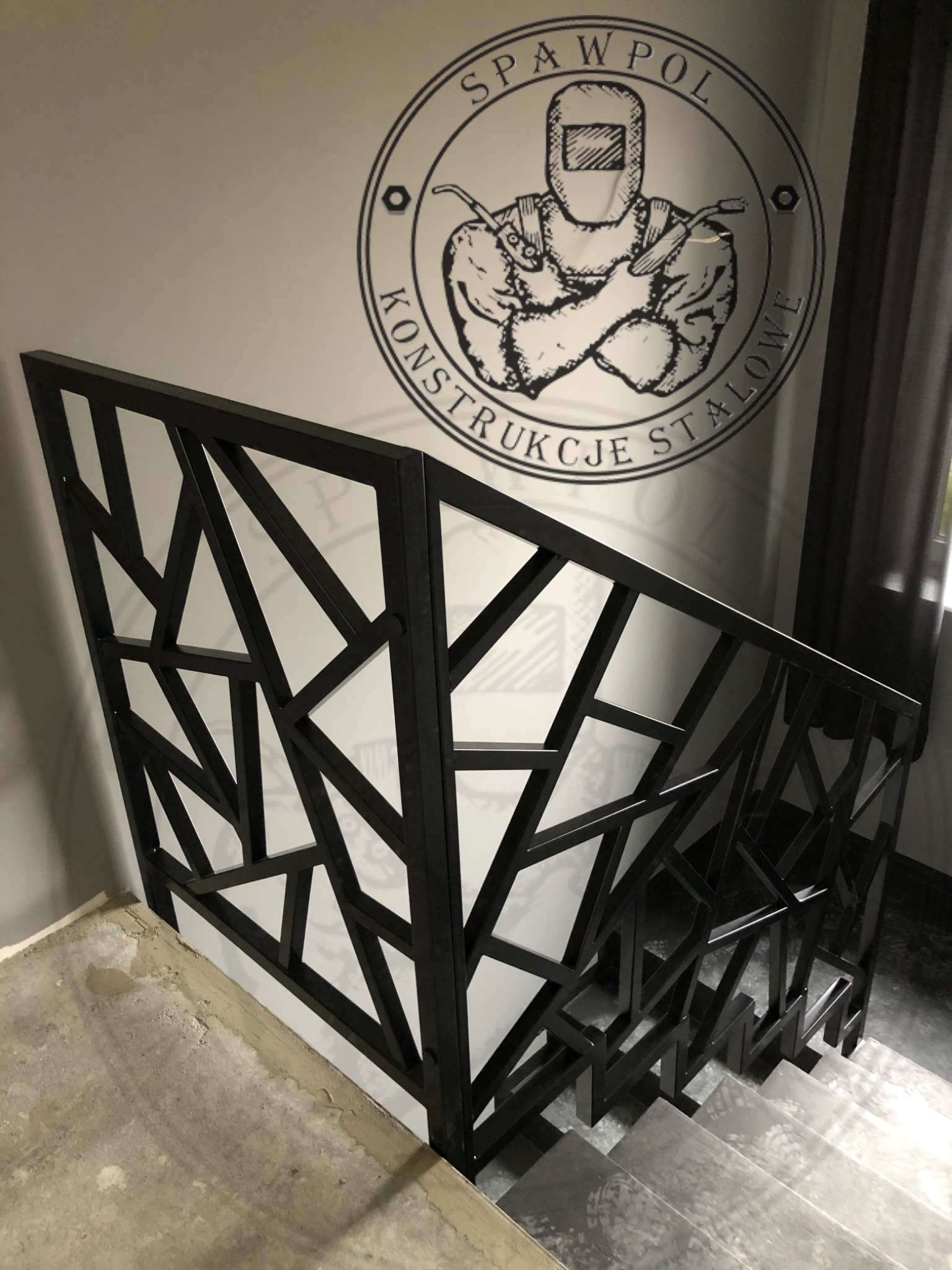 Nowoczesna balustrada schodowa z czarnego metalu o geometrycznym wzorze, widoczna z góry, z logo firmy 'SPAWPOL KONSTRUKCJE STALOWE' na ścianie w tle.