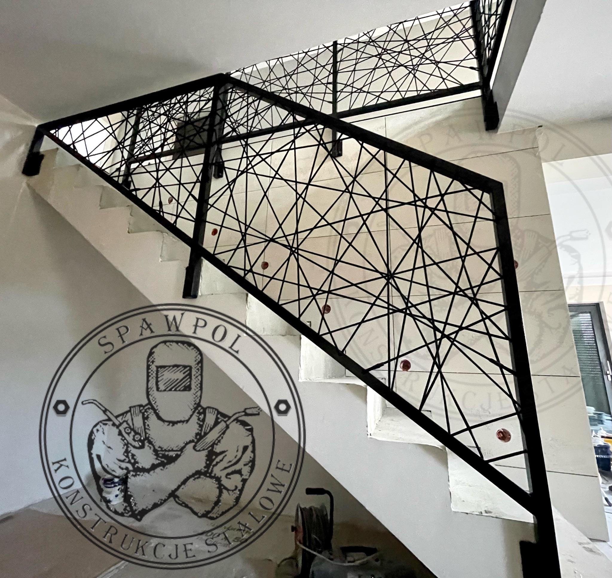 Nowoczesna balustrada schodowa z czarnego metalu o geometrycznym wzorze, zamontowana na białych schodach w jasnym wnętrzu. Widoczny fragment logo firmy.