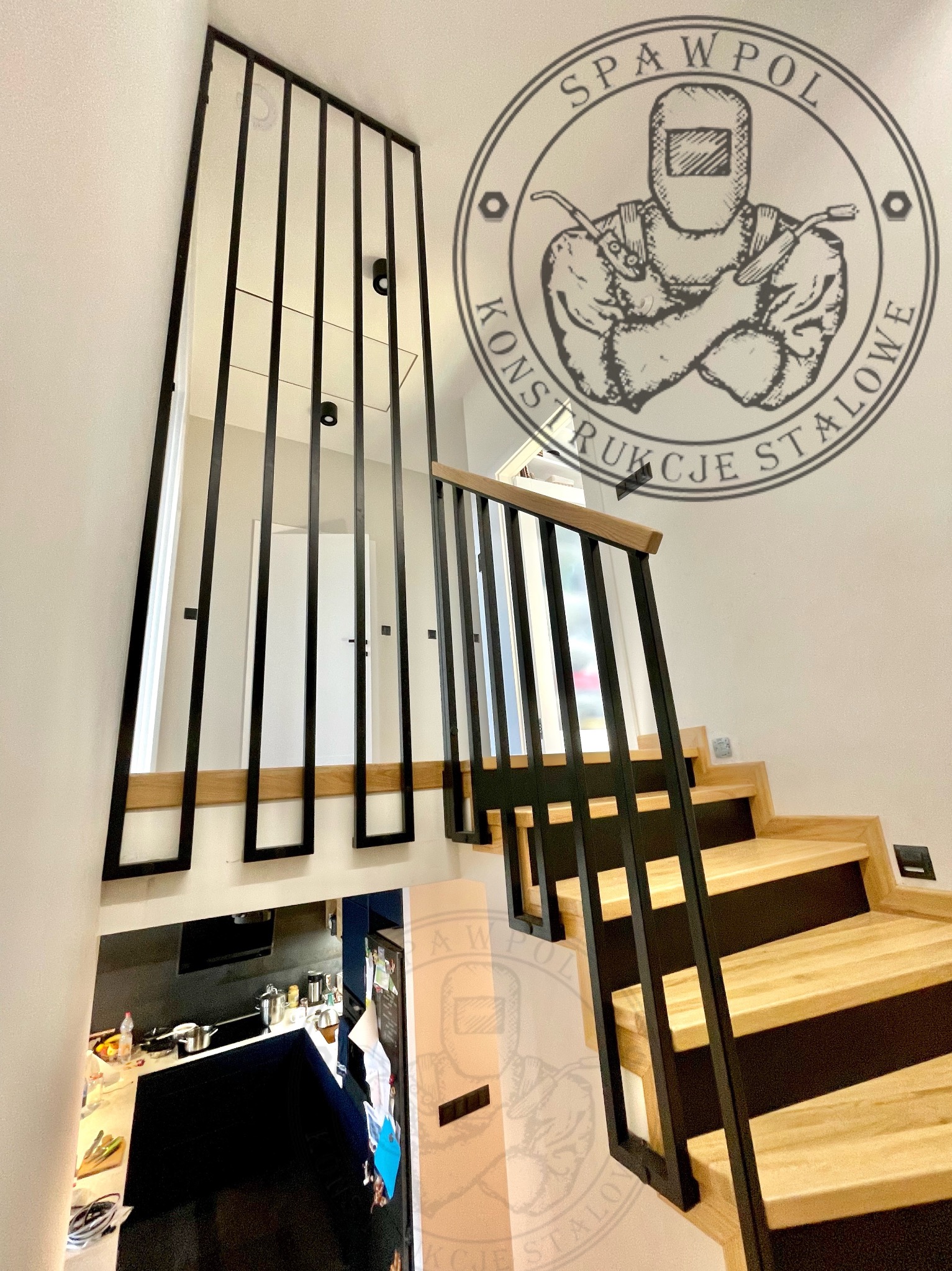 Nowoczesne schody z drewnianymi stopniami i czarną, metalową balustradą z pionowymi szczeblami. Widok z dołu, logo firmy na ścianie.