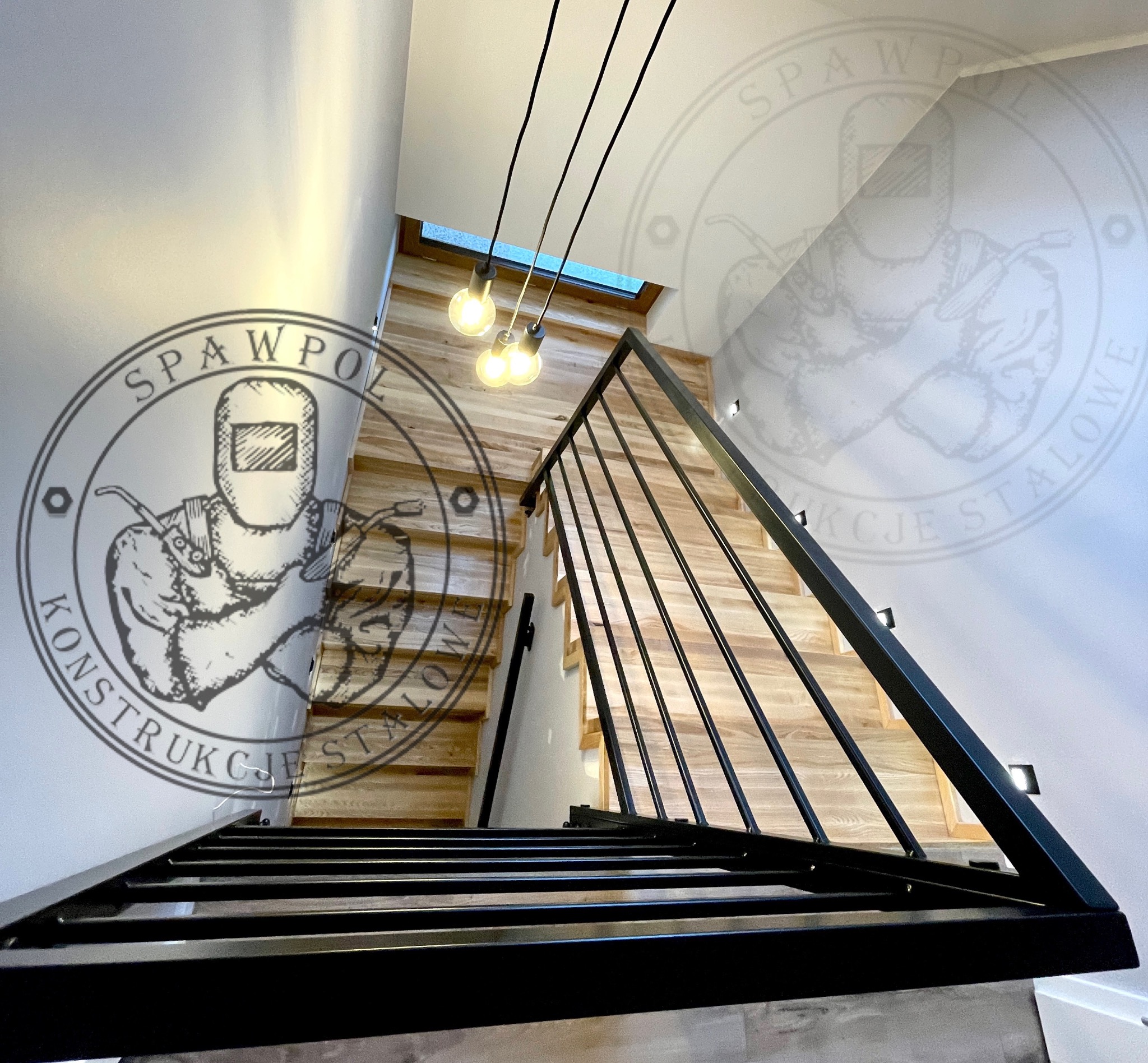 Nowoczesne schody z drewnianymi stopniami i czarną metalową balustradą, oświetlone wiszącymi lampami. Widok z dołu, z logo firmy na ścianie.