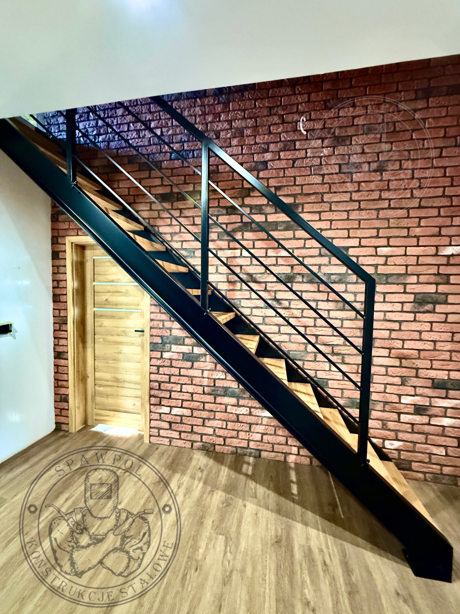 Nowoczesne schody metalowe z drewnianymi stopniami, czarna konstrukcja, ceglana ściana w tle, widoczne logo firmy na podłodze. Minimalistyczny design.