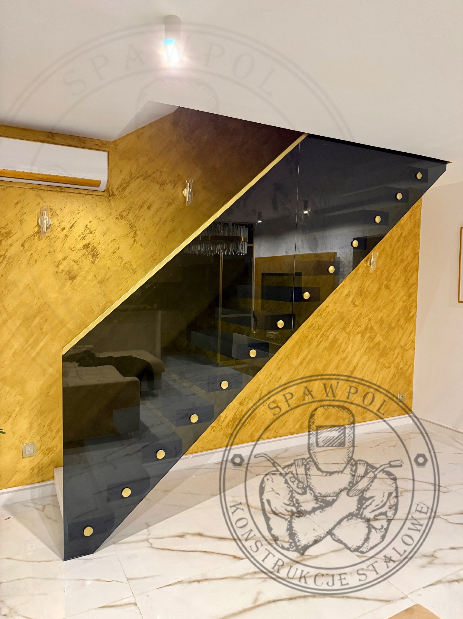 Nowoczesne schody z balustradą szklaną, złote akcenty na stopniach i ścianie, marmurowa podłoga. Widok na salon przez szklaną balustradę.