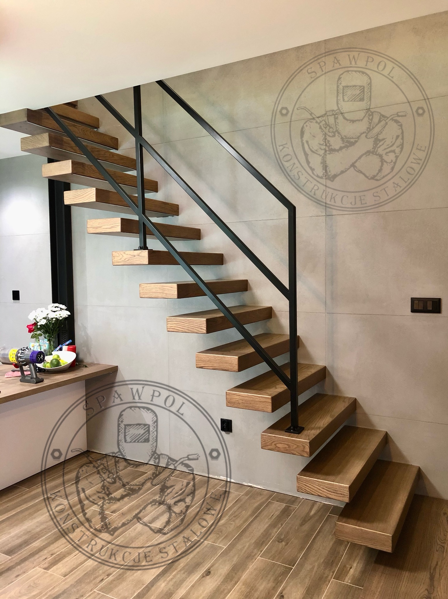 Nowoczesne schody wiszące z drewnianymi stopniami i czarną metalową balustradą, zamontowane w jasnym wnętrzu z szarą ścianą i drewnianą podłogą.