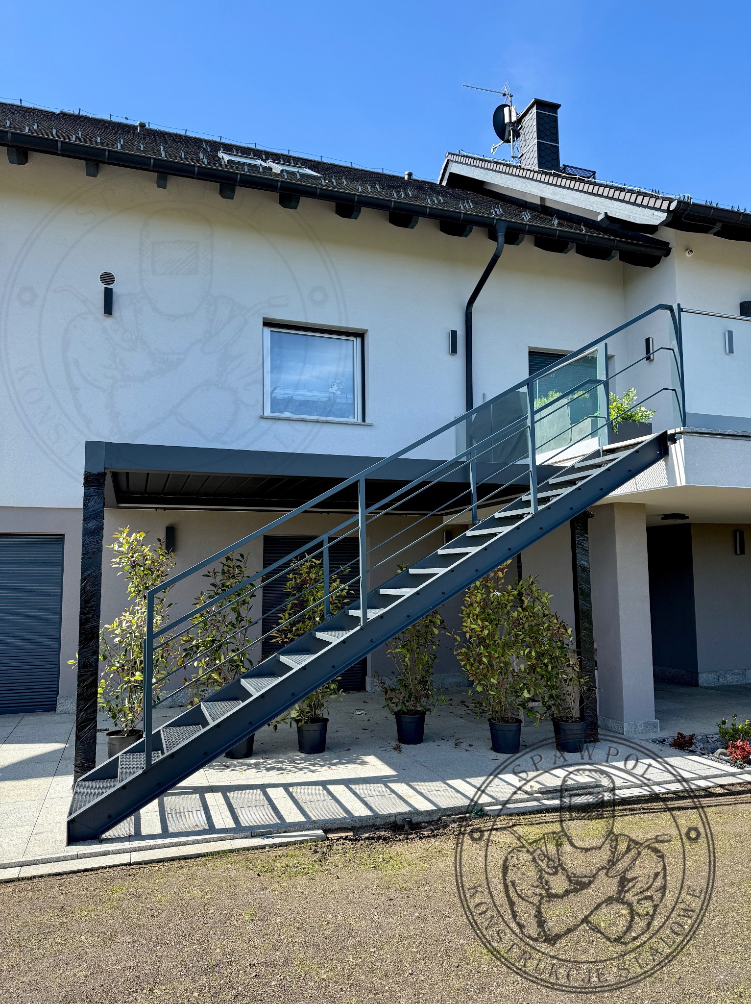 Zewnętrzne schody metalowe z balustradą szklaną prowadzące do wejścia domu, z roślinami w donicach po bokach. Widok na elewację budynku w słoneczny dzień.