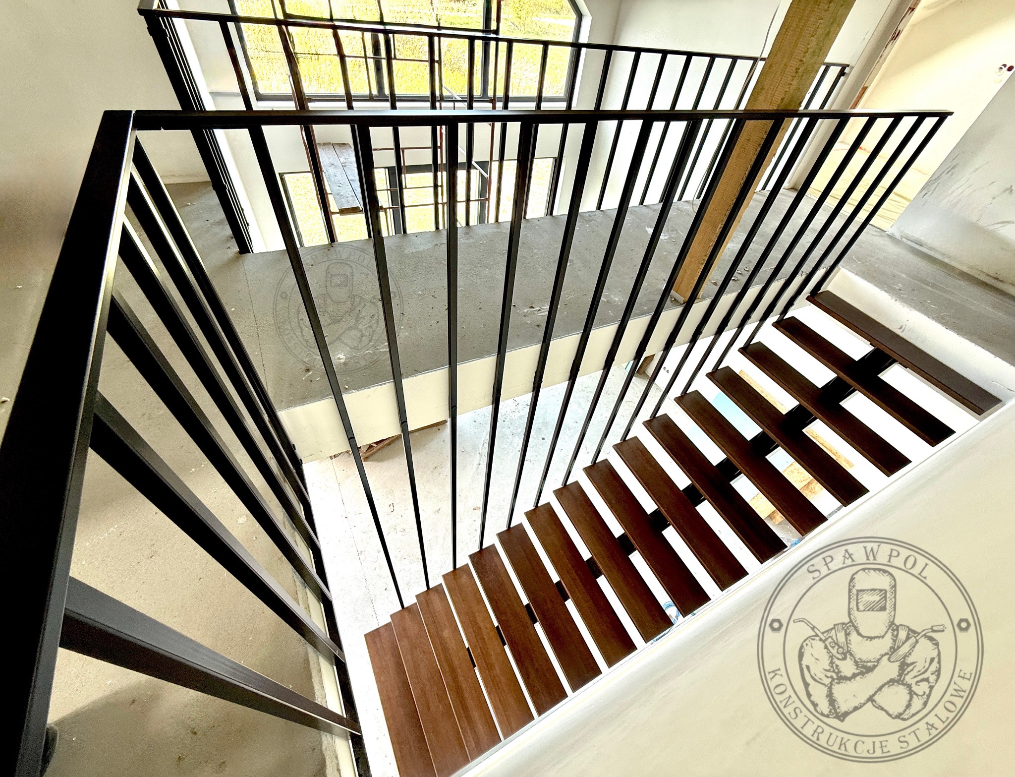 Nowoczesne schody z drewnianymi stopniami i czarną metalową balustradą, widok z góry, logo firmy w rogu. Minimalistyczny design, surowe wnętrze w trakcie wykończenia.