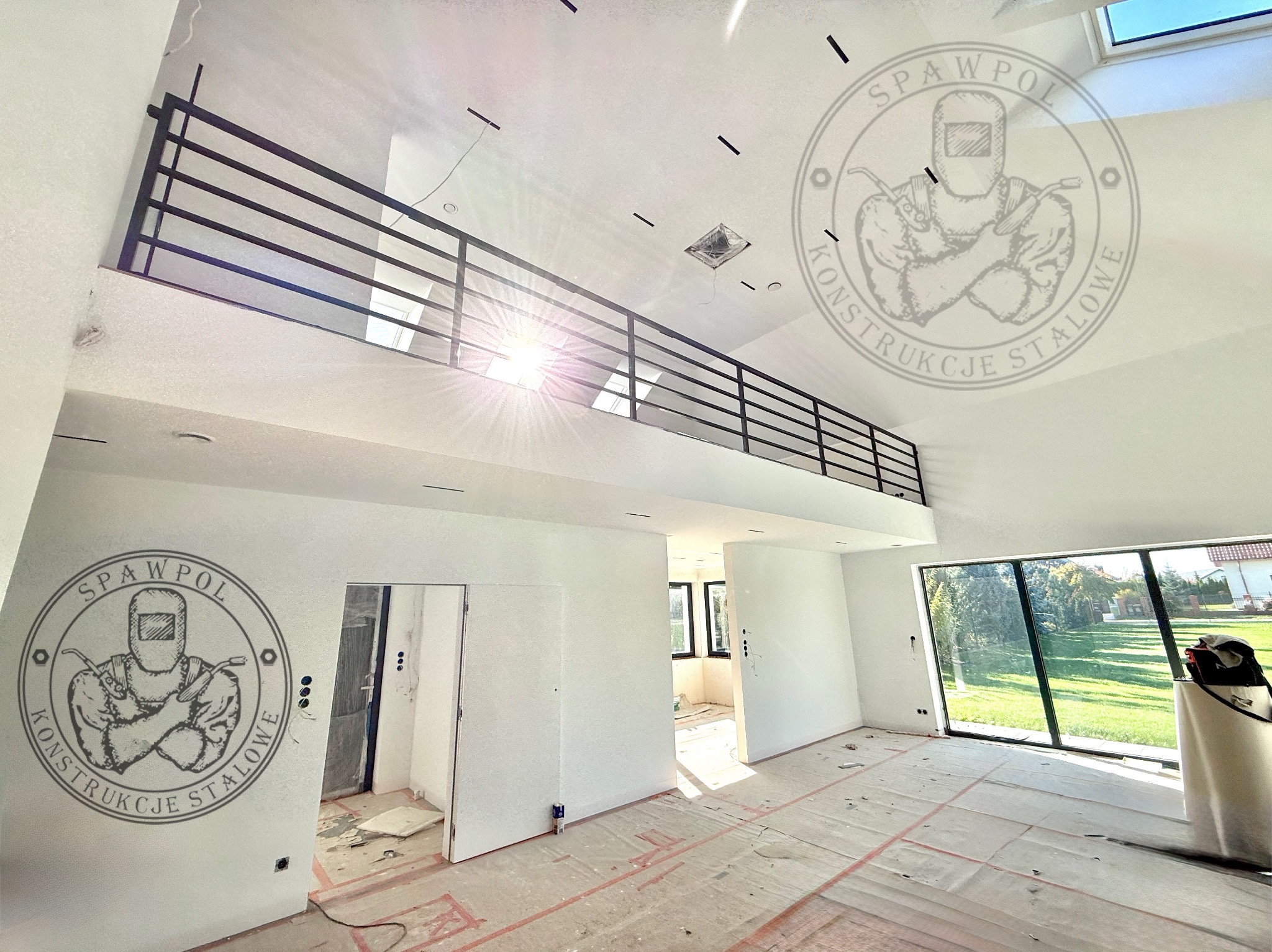 Wnętrze domu z czarną, metalową balustradą na antresoli, widok z dołu. Duże okno z widokiem na zewnątrz. Na ścianie logo firmy. Podłoga zabezpieczona folią.