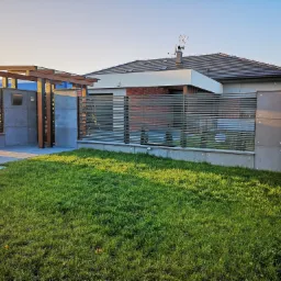 Nowoczesne ogrodzenie posesji z poziomymi przęsłami, betonowymi słupkami i elementami drewnianymi, widok z zewnątrz na tle domu z szarą dach&oacute;wką i zielonym trawnikiem.