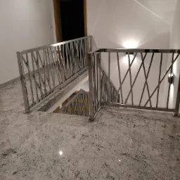 Wnętrze klatki schodowej z balustradą ze stali nierdzewnej o geometrycznym wzorze, widok z góry na schody wykończone płytami imitującymi kamień.