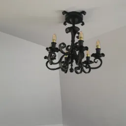 Czarny, kuty żyrandol sufitowy z czterema żarówkami w kształcie płomienia, zamontowany na białym suficie.