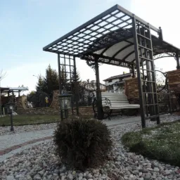 Kuta altana z siedziskiem i drabinką w ogrodzie, z dekoracyjnym ogrodzeniem z kamienia i metalu w tle, na ścieżce z kamieni.