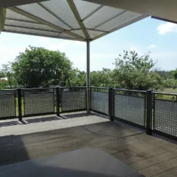 Zadaszony taras z balustradą o ażurowym wypełnieniu, widok na zieleń i zabudowania.