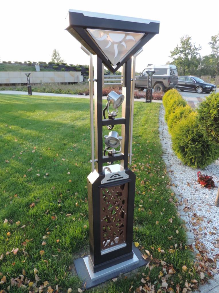 Nowoczesna lampa ogrodowa z metalu i szkła z motywem słońca, oświetlająca trawnik i ścieżkę żwirową w parku.