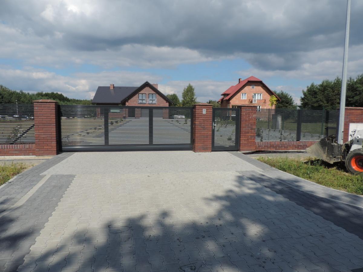 Automatyczna brama przesuwna z poziomych profili, osadzona między ceglanymi słupkami, na tle dwóch domów i pochmurnego nieba. Widoczny fragment maszyny budowlanej.