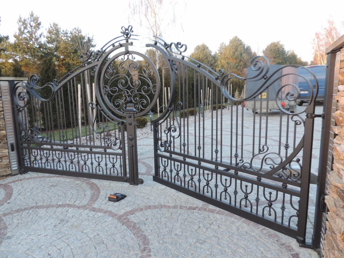 Otwarta, dwuskrzydłowa brama z kutego żelaza z dekoracyjnymi elementami, na podjeździe wyłożonym kostką brukową, z widocznym samochodem dostawczym w tle.