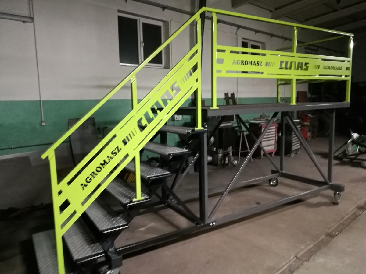 Metalowa platforma robocza z schodami, pomalowana na jaskrawy zielony kolor, z logo firmy Agromasz-Claas, na kółkach, w warsztacie.