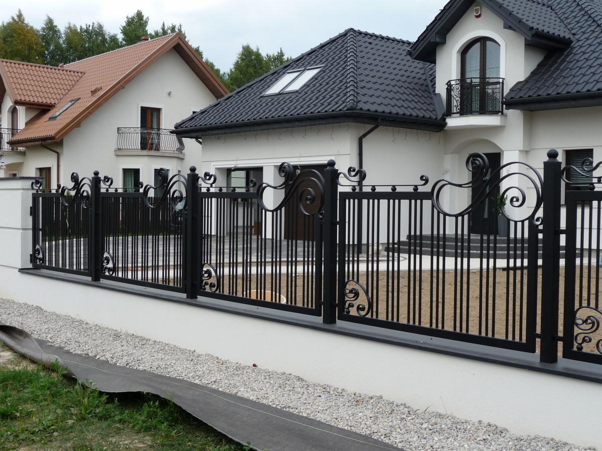 Czarny, metalowy płot z dekoracyjnymi, kutymi elementami, oddzielający posesję z dwoma domami o białych elewacjach i ciemnych dachach, widok z zewnątrz.