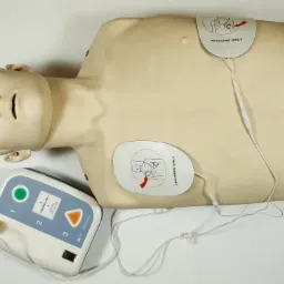 Manekin do nauki pierwszej pomocy z podłączonym defibrylatorem treningowym i elektrodami, oznaczonymi napisem 'TRAINING ONLY'.