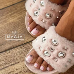 Paznokcie u stóp z białym francuskim manicure i drobnymi kryształkami na dużym palcu, prezentowane w różowych, futrzanych klapkach z metalowymi ozdobami na drewnianej podłodze.