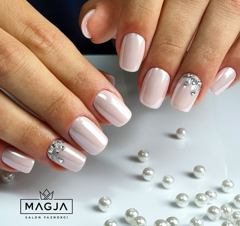 Delikatny, perłowy manicure w odcieniach nude, ozdobiony drobnymi kryształkami na jednym paznokciu, wykonany w salonie w Krakowie. Drobne perły rozrzucone wokół dłoni dodają elegancji.