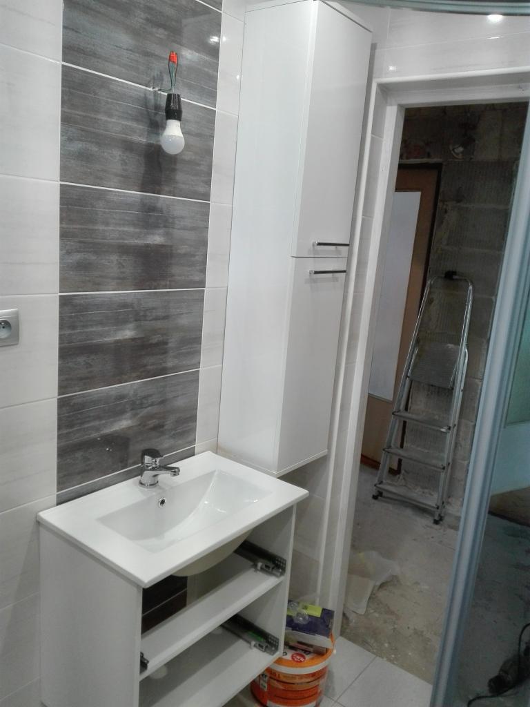 Wykończona łazienka z szarymi płytkami imitującymi drewno, białą szafką nad umywalką i wiszącą żarówką bez klosza, widoczna drabina w przejściu do kolejnego pomieszczenia.