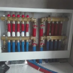 Rozdzielacz ogrzewania podłogowego w białej obudowie, z widocznymi rurami w kolorach czerwonym i niebieskim, zamontowany na tle surowej ściany.