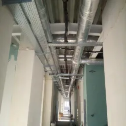 Długi korytarz w budynku w trakcie instalacji systemu wentylacyjnego z widocznymi kanałami wentylacyjnymi i korytkami kablowymi pod sufitem.