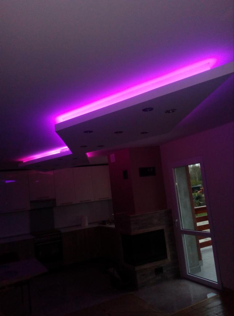 Nowoczesny salon z kuchnią, oświetlenie sufitowe LED w kolorze fioletowym, podwieszany sufit z punktowymi lampami, widok na kominek i wyjście na balkon.