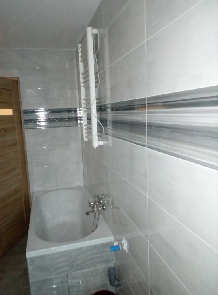 Wykończona łazienka w szaro-białych płytkach z dekoracyjnym pasem, biała wanna z baterią i grzejnik drabinkowy na ścianie, widoczne elementy instalacji wodnej.