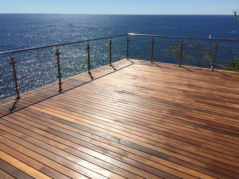 Wykończony drewniany taras z widokiem na morze, chromowane barierki z przezroczystym szkłem, różne odcienie drewna na desce tarasowej.