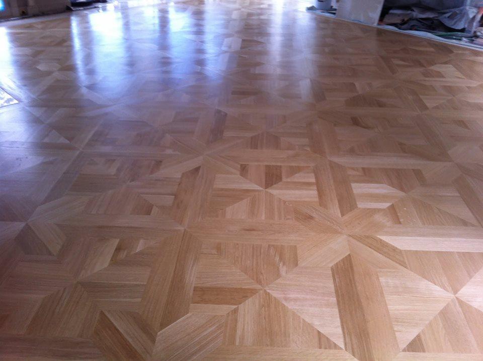 Wypolerowany parkiet ułożony we wzór geometryczny z widocznymi refleksami światła, ukazujący różnice w odcieniach drewna.