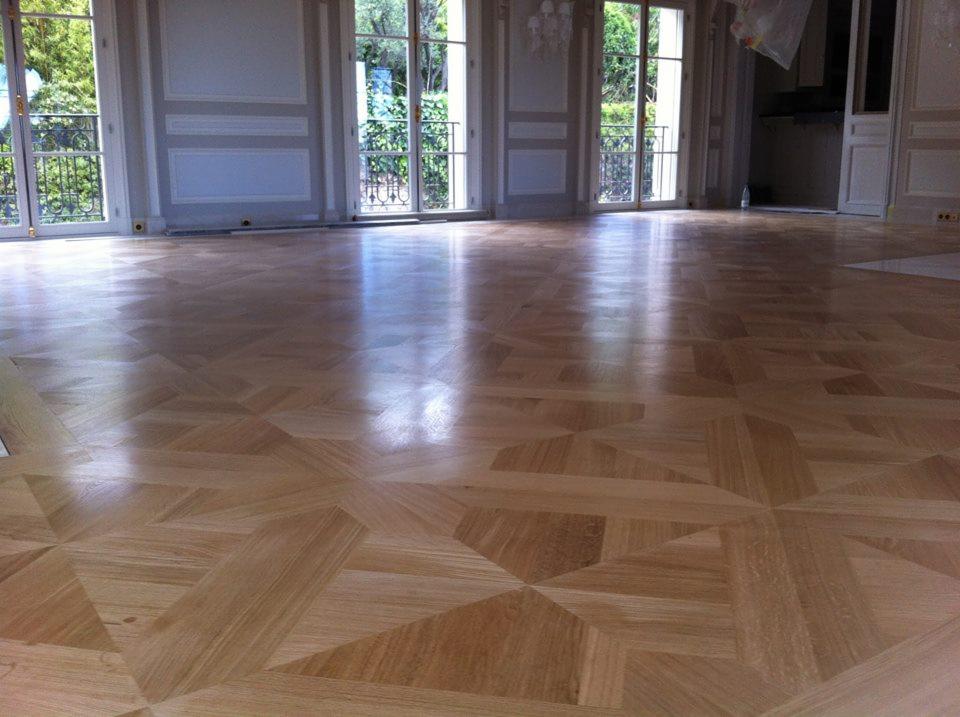 Jasna, lakierowana podłoga z parkietu o geometrycznym wzorze w przestronnym, jasnym wnętrzu z wysokimi oknami balkonowymi i sztukateriami.