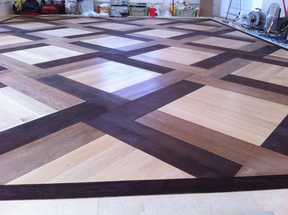 Ułożona podłoga z parkietu w geometryczny wzór z wykorzystaniem drewna w różnych odcieniach brązu, z widocznymi narzędziami i materiałami budowlanymi w tle.