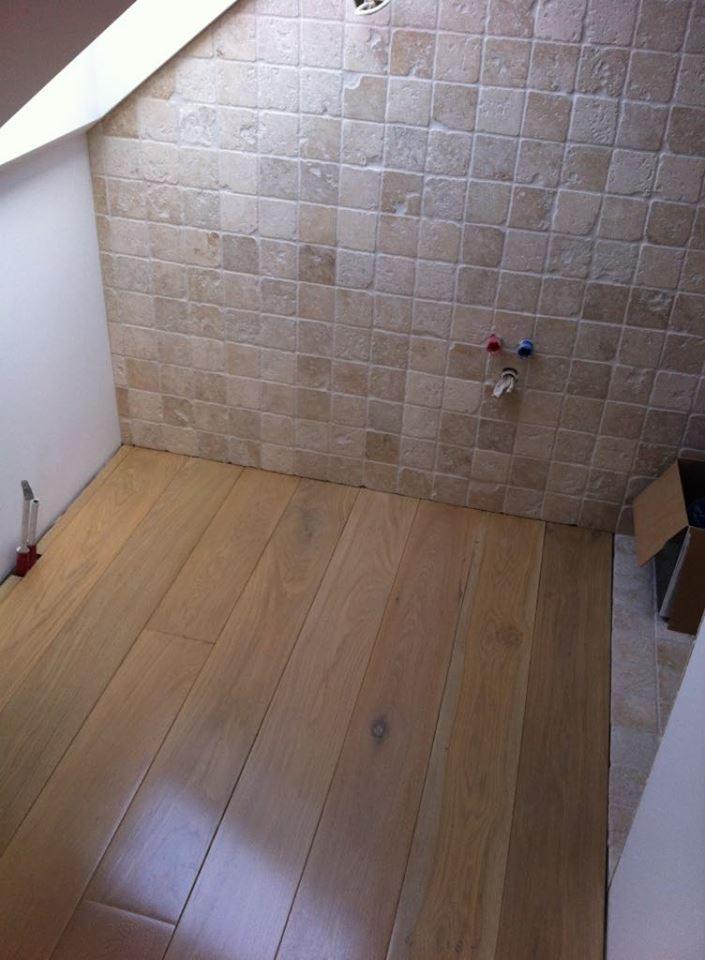 Układanie jasnego parkietu w łazience z beżowymi płytkami na ścianie, widoczne wystające rury wodne i narzędzie do układania parkietu przy ścianie.
