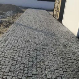 Ułożona nawierzchnia z szarej kostki granitowej, widok z perspektywy, z fragmentem kontenera po prawej i stertą piasku po lewej stronie.