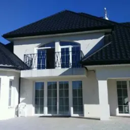 Biały dom z czarnym dachem i balkonem z ciemnymi barierkami, widok z dołu na tle błękitnego nieba.