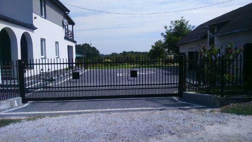 Czarna, przesuwna brama wjazdowa z pionowych prętów, prowadząca na posesję z dwoma budynkami w tle, widok z poziomu żwiru.