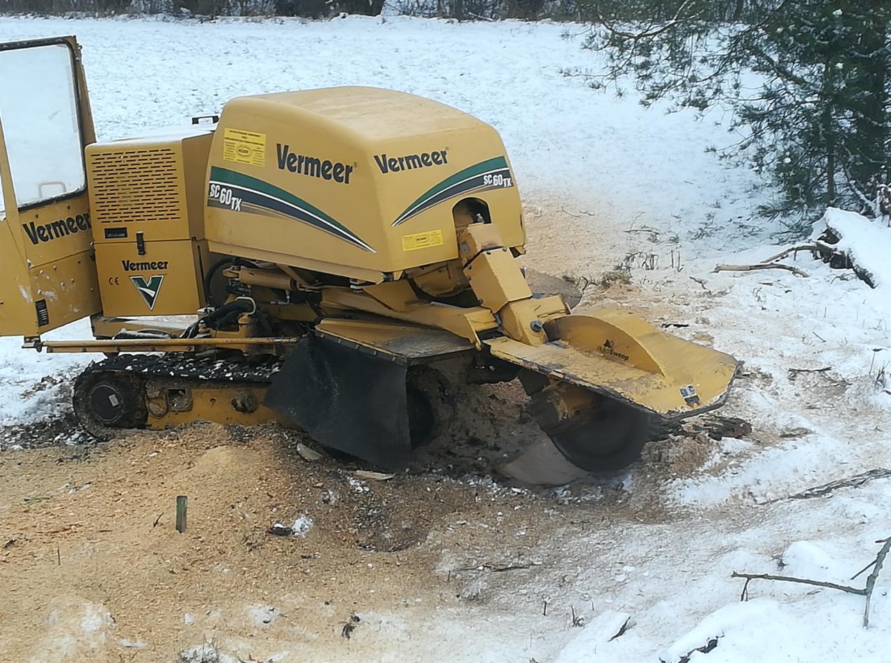 Żółta frezarka do pni drzew marki Vermeer SC60TX na gąsienicach, z widocznymi trocinami i resztkami po frezowaniu pnia, zimowy krajobraz w tle.