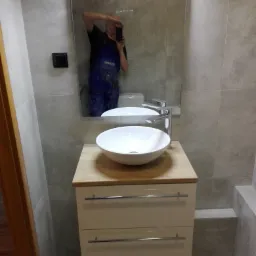 Nowoczesna łazienka z okrągłą umywalką nablatową, drewnianym blatem i szafką z dwoma szufladami, widoczna w lustrze osoba robiąca zdjęcie.