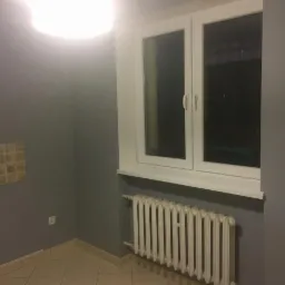 Szare wnętrze pokoju po malowaniu, widoczne okno PCV, biały grzejnik żeberkowy i fragment płytek na ścianie, oświetlone górnym światłem.