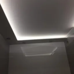 Biały sufit z oświetleniem LED wzdłuż krawędzi i punktowymi reflektorami, odbicie światła na błyszczącej ścianie.