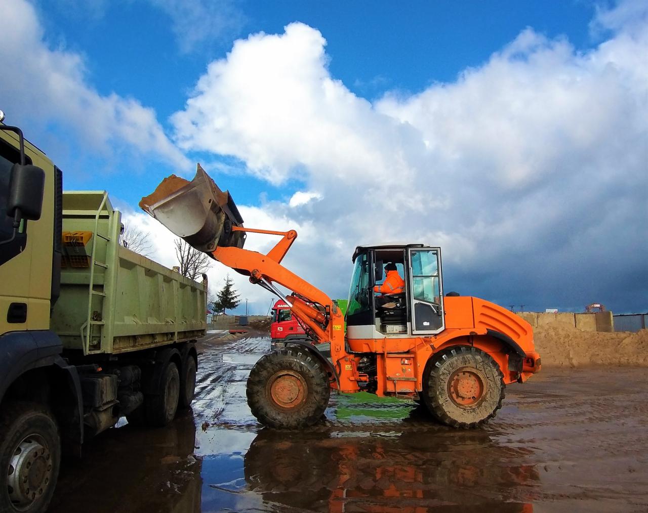 Pomarańczowa ładowarka kołowa z pełną łyżką ziemi podnosi ją nad zieloną ciężarówką na placu budowy w pochmurny dzień.