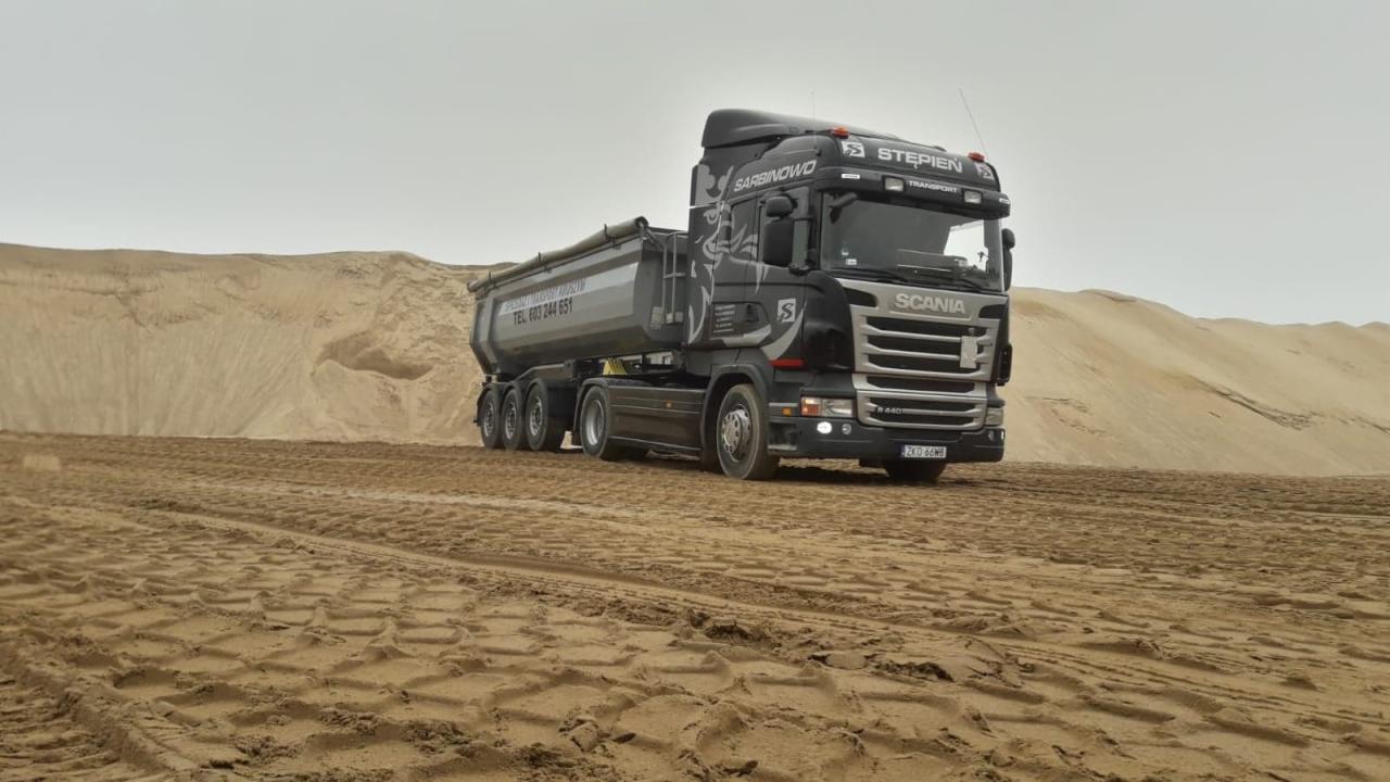 Czarna ciężarówka Scania z naczepą wywrotką firmy Stepien Transport Sarbinowo na piaszczystym terenie z widocznymi śladami opon.