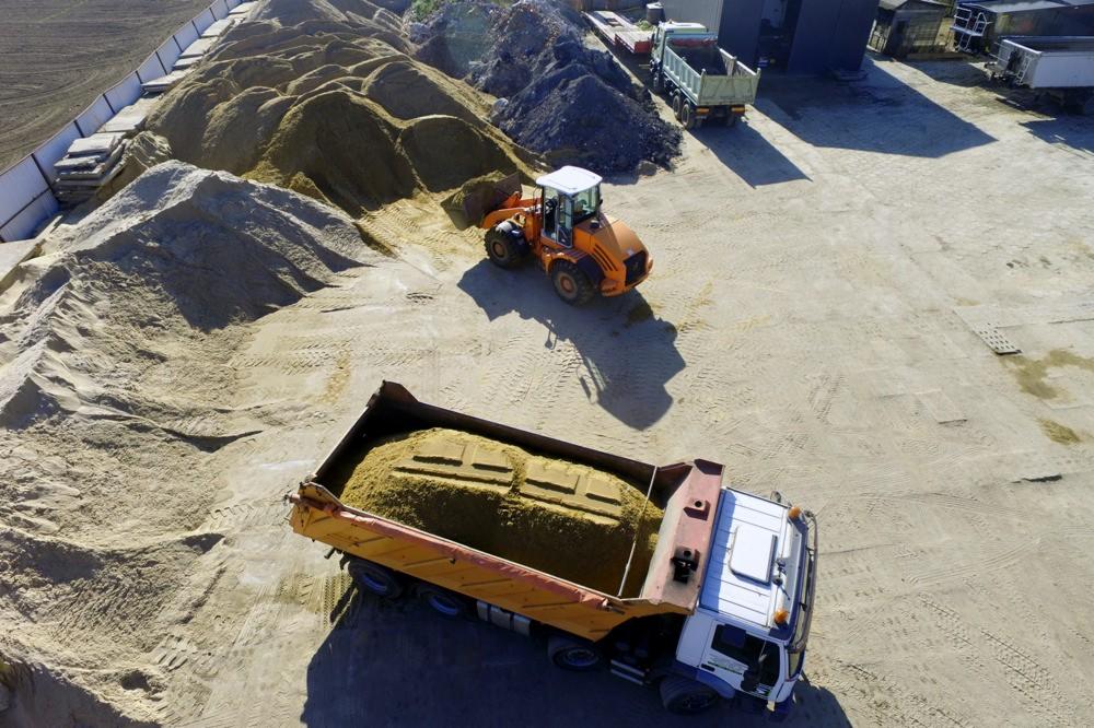 Pomarańczowa ładowarka kołowa nabiera piasek i ładuje go na ciężarówkę z otwartą skrzynią ładunkową; widok z góry na plac budowy z hałdami piasku i innymi pojazdami.