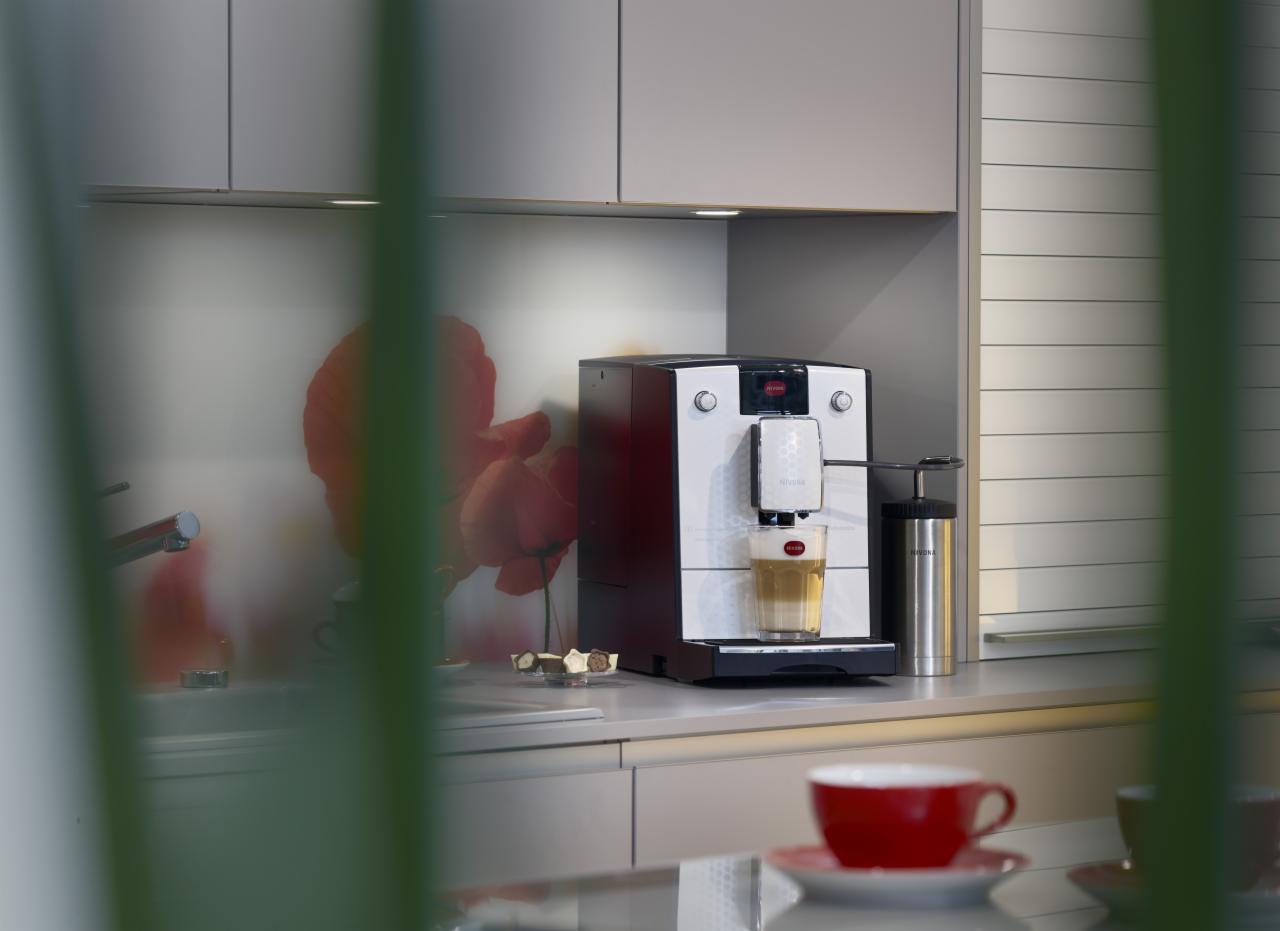 Automatyczny ekspres do kawy Nivona w nowoczesnej kuchni z motywem czerwonych maków na ścianie, przygotowujący latte macchiato, obok stalowy pojemnik na mleko i miseczka z czekoladkami.