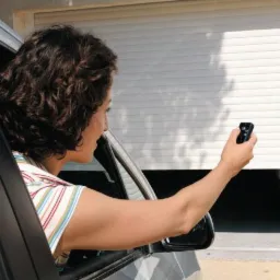 Kobieta z samochodu pilotem otwiera białą, poziomą osłonę okienną garażu.