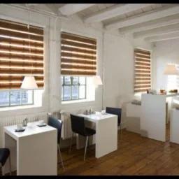 Jasne wnętrze kawiarni z trzema oknami przesłoniętymi poziomymi przesłonami w odcieniach brązu, białe stoliki i krzesła, drewniana podłoga, grzejniki pod oknami.