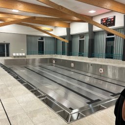 SOLIDTRANS-TEAM ILONA ARCISZEWSKA - Opróżniony basen sportowy z metalowym dnem i drewnianą konstrukcją dachu. Widoczne okna, płytki podłogowe i tablica wyników.