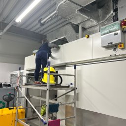 SOLIDTRANS-TEAM ILONA ARCISZEWSKA - Pracownik na rusztowaniu czyści białe panele w hali przemysłowej. Widoczny żółty odkurzacz przemysłowy i elementy wentylacji pod sufitem.